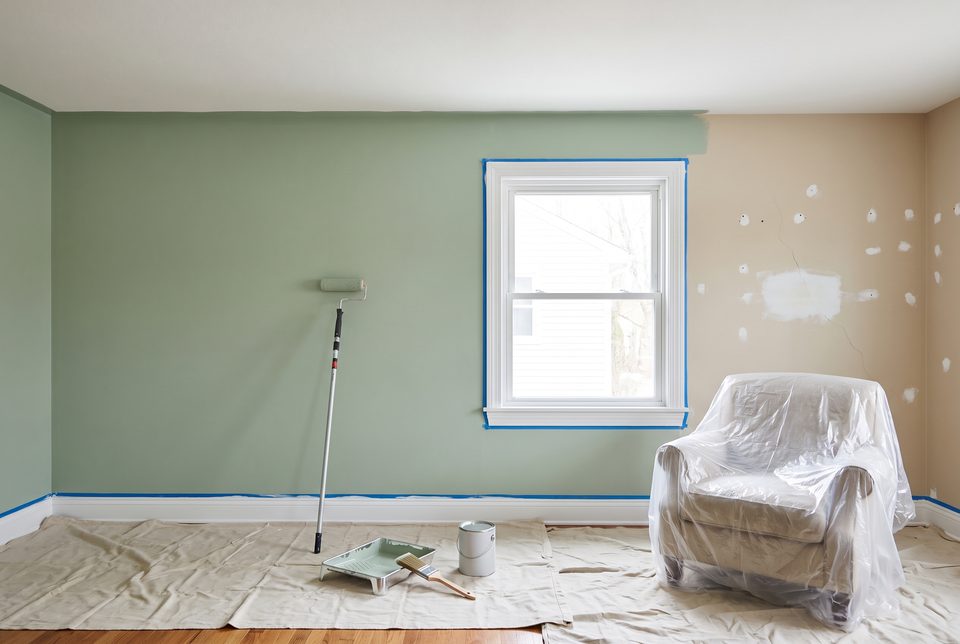 A living room part-way through a repaint: sage green rolled over most of the wall with a clean line at the ceiling, patched nail holes and a sanded crack on the old beige section, taped window trim, drop cloths on the floor and an armchair wrapped in plastic